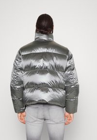 Person med langt mørkt hår i en knold, iført en skinnende grå dunjakke og lysegrå jeans, står med ryggen til en simpel hvid baggrund.