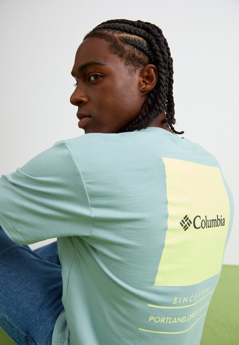 T-shirt bleu clair à manches courtes avec un design carré jaune au dos, comportant le logo Columbia et des détails textuels. Matière coton.