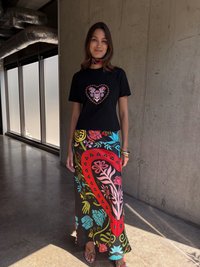 Μαύρη μπλούζα t-shirt με γραφικό καρδιάς, πολύχρωμη φλοράλ φούστα μάξι με σχέδια σε κόκκινο, μπλε και ροζ, και λουράτα καφέ σανδάλια.