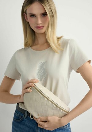 Kobieta o blond włosach do ramion nosi jasnobeżową koszulkę z motywem pingwina z cyrkoniami, trzyma pikowaną kremową torbę na zamek.