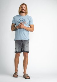 Ljust blå bomullströja med tryckt grafik, kombinerad med slitna grå denim shorts och bruna sandaler. En enkel, avslappnad sommaroutfit.