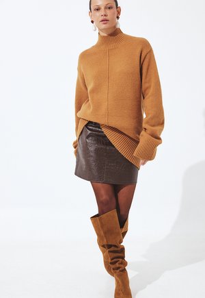 Femme portant un pull à col roulé camel, une mini-jupe texturée marron foncé, des collants transparents et des bottes souples en daim camel montant jusqu'aux genoux.