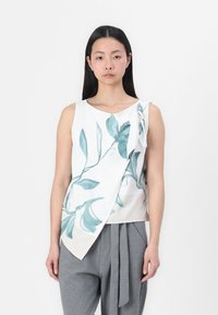 Haut sans manches blanc avec un imprimé botanique vert clair, présentant un design asymétrique et un détail noué à l'épaule. Associé à un pantalon gris.