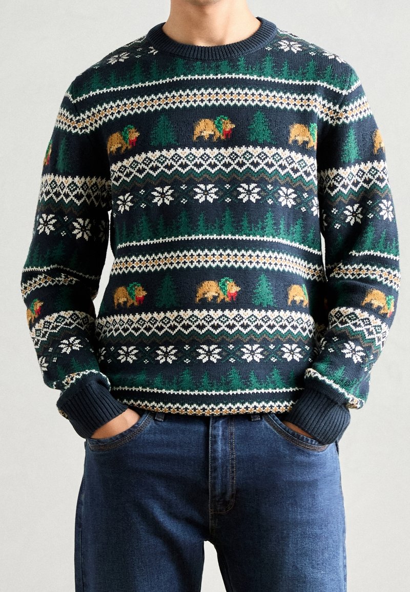 Homme portant un pull de Noël bleu marine avec des motifs d'ours, d'arbres verts et de flocons de neige blancs, associé à un jean bleu.