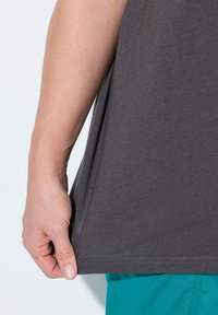 Graues Kurzarmhemd mit glatter Textur, abgerundetem Saum und seitlichen Schlitzen, kombiniert mit türkisfarbenen Shorts, die unten sichtbar sind.