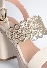 Beige blokhak sandaal met een brede band met gouden ronde accenten en transparante ringen. Gladde textuur met een roze binnenzool.