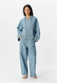 Denim-Outfit mit einer blauen Kapuzenjacke mit Vorderstaschen und weiten Hosen. Glatte Textur mit sichtbaren Nähdetails.