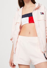 Jasnoróżowa kurtka na zamek z kapturem, zestawiona z granatowo-czerwono-białym crop topem w paski oraz dopasowanymi różowymi szortami. Miękki materiał bawełniany.