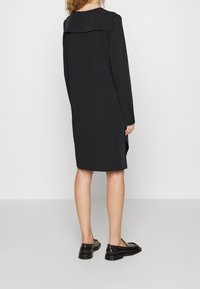 Mustapaitainen pitkähihaisessa mekossa on taitettu kaulus, polvipituinen ja sivurei'illä varustettu. Yhdistetty mustiin loafereihin, jotka korostavat sujuvaa ja sileää tekstuuria.