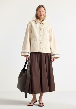 Kvinde iført en cremefarvet jakke med vide ærmer, en lang brun nederdel, bærende på en stor mørk tote-taske og iført brune sandaler med hæl og remme.