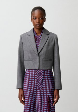 Donna che indossa un blazer grigio corto sopra un vestito viola con motivo circolare blu, in piedi davanti a uno sfondo chiaro uniforme.