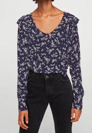 Femme portant un chemisier bleu marine à manches longues avec un petit motif floral blanc et un jean noir taille haute.