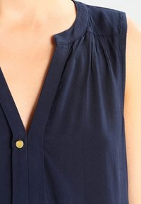 Mörkblå ärmlös blus med en liten krage och en knappdetalj, med mjuka veck längs axelpartiet.