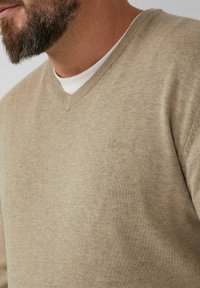 Beige v-hals trui met een fijne breistekstuur, voorzien van een subtiele merkl logo op de borst. Gemaakt over een wit shirt.