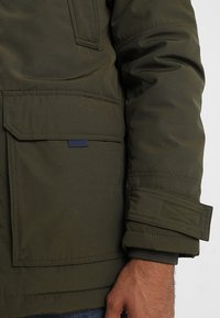 Olivgrön, vadderad jacka med en stor framficka, sydda detaljer och manschettärmar. Hållbart tyg med en slät textur.