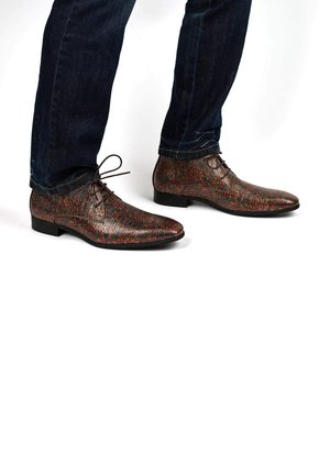 Braune gemusterte Lederschuhe mit spitzer Zehenform, Schnürdesign und niedrigem Absatz, getragen mit dunkelblauen Jeans. Strukturierte Oberfläche mit mehrfarbigen Akzenten.
