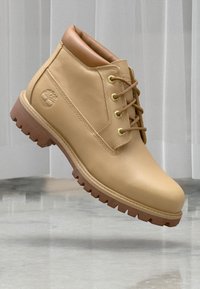 Beige skinnankelstøvel med såle i brunt gummi, rund tå og beige snøring. Har en logo på siden og en polstret krage.