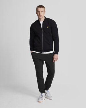 Black zip-up sweatshirt with a yellow logo, paired with dark jogger pants and grey sneakers. Plain white t-shirt underneath.