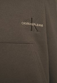 Sudadera de Calvin Klein Jeans en tejido gris oscuro que presenta un logo bordado en negro con texto en blanco. Textura suave e incluye un bolsillo tipo canguro.