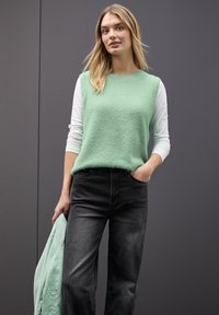 Miętowa, puszysta bluzka bez rękawów na białej koszuli z długim rękawem, w połączeniu z czarnymi dżinsami. Gładka tekstura, luźny krój.