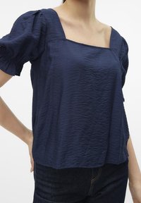 Mörkblå, strukturerad topp med fyrkantig halsringning och puffärmar, gjord av lättviktsmaterial. Har en avslappnad passform och lätt lyster.