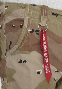 Pantalones cortos de cargo en camuflaje de tejido con patrones en marrón, verde y negro; cuenta con un anillo de metal y una etiqueta roja que dice "RETIRE ANTES DEL VUELO".