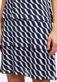 Marineblaues Kleid mit weißem Wellenmuster, geschichteter Gestaltung, fließendem Stoff und abschließendem Saum. Hat einen lockeren Sitz an Taille und Hüften.