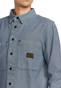 Camisa de mezclilla azul claro con cuello vuelto, botones negros y un bolsillo en el pecho izquierdo que lleva una etiqueta de marca.