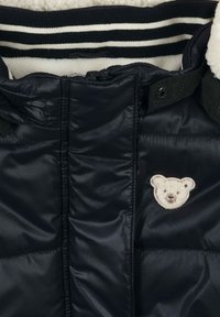 Schwarze Daunenjacke mit einem weichen, Fleece-Futter. Sie hat ein Bärenapplikation auf der Brust und einen gerippten, gestreiften Kragen.