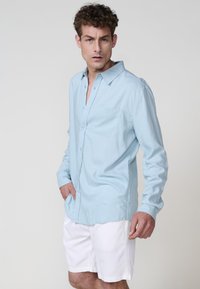 Camicia a maniche lunghe azzurro chiaro in tessuto morbido, con design a bottoni e taschino sul petto. Indossata con pantaloni corti bianchi.