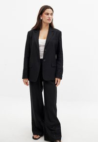 Black blazer with a tailored fit, featuring a single button closure and detailed embroidered cuffs. Paired with wide-leg black trousers.