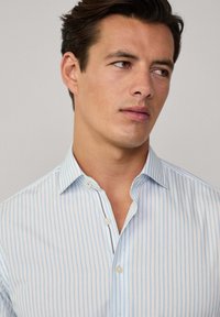 Chemise boutonnée à rayures bleu clair et blanche, avec un col classique et une patte de boutonnage, tissu lisse et détails subtils.