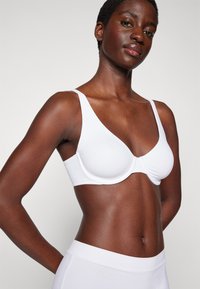 Soutien-gorge blanc rembourré avec de larges bretelles et une forme en V profonde ; tissu lisse, design minimaliste ; associé à un bas blanc taille haute.