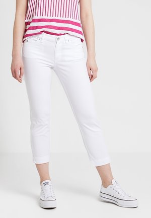 Frau trägt weiße kurze Jeans, rot-weiß gestreiftes Oberteil und weiße Sneaker mit niedrigem Schaft vor einfarbigem Hintergrund.