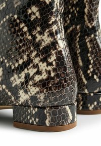 Primer plano de botas de tobillo con estampado de serpiente en tonos marrón y beige, con textura hexagonal y tacones bajos en un fondo blanco.