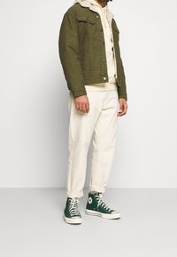 Giacca di velluto a coste verde oliva con colletto in pelliccia sintetica crema, indossata sopra un hoodies beige, abbinata a pantaloni a sigaretta crema e sneakers alte verdi.