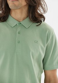 Pikétröja i ljusgrön, tillverkad av bomull, med krage, tre knappar och en liten logotyp på vänster sida. Slät textur.