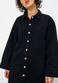Schwarzes Denim-Hemdkleid mit Kragen, zwei Brusttaschen und silbernen Knöpfen, die vorne entlanglaufen. Lockerer Schnitt und lange Ärmel.