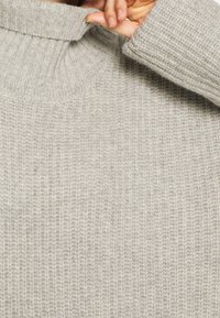 Pull en col roulé à côtes de couleur gris clair, avec un col haut et des manches longues. La texture est douce et confortable, idéale pour le superposition.