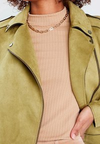 Krøllet hår person iført en beige struktureret sweater, guld kæde halskæde og lysegrøn ruskind jakke med metal knapper og lynlås.