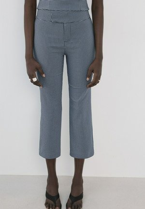 Person iført korte sort-hvide ternede bukser, sorte åbne sandaler og sølvringe på begge hænder, stående op ad en ensfarvet væg.