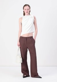 Hvit ermeløs topp med vridd knutetail kombinert med brune vidde bukser. Modellen holder en liten beige håndveske; har på seg svarte spisse sko.