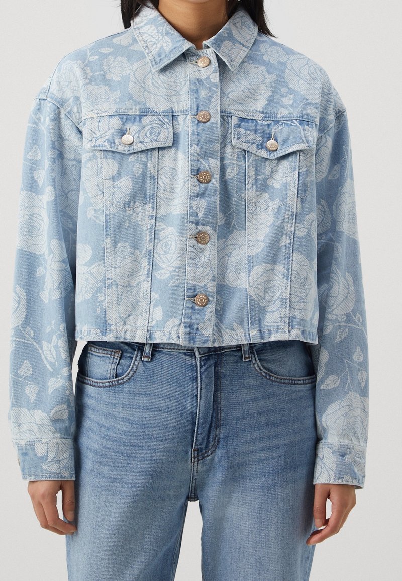 Femme portant une veste en jean courte bleu clair avec motif floral blanc, boutons argentés et jean bleu taille haute.