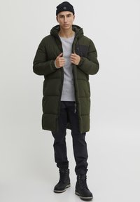 Oliven grøn pufferjakke med en sort sidelomme, hætte og elastiske manchetter, båret over en grå sweatshirt og mørke bukser, med sorte støvler.