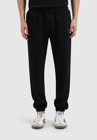 Zwarte joggingbroek met een elastische tailleband en taps toelopende boorden. Bevat een subtiel logo nabij de heup. Glad, zacht materiaal met een ontspannen pasvorm.