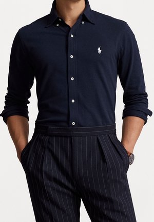Homme portant une chemise bleu marine boutonnée avec un logo blanc et un pantalon à fines rayures foncé, les mains dans les poches et une montre au poignet gauche.