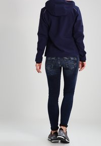 Marineblaues Jacket mit Kapuze, taillierter Schnitt, kombiniert mit dunkelblauen engen Jeans. Die Schuhe sind grau mit schwarzen Akzenten. Sichtbare Nähte an den Jeans.