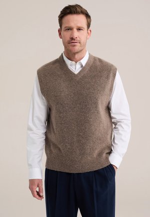 Homme portant un gilet en laine marron sur une chemise blanche et un pantalon bleu foncé, debout devant un fond clair.