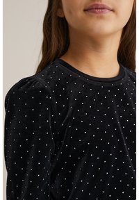 Fekete bársony felső ezüst pöttyös részletekkel, kerek nyakkivágással és puffos ujjakkal. A textúrája sima és puha, diszkrét csillogást kölcsönöz.