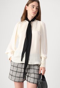 Elfenbensfarget bluse med blonder detaljer, lange ermer og et svart slips. Kombinert med grå tweed-shorts med rutete mønster.